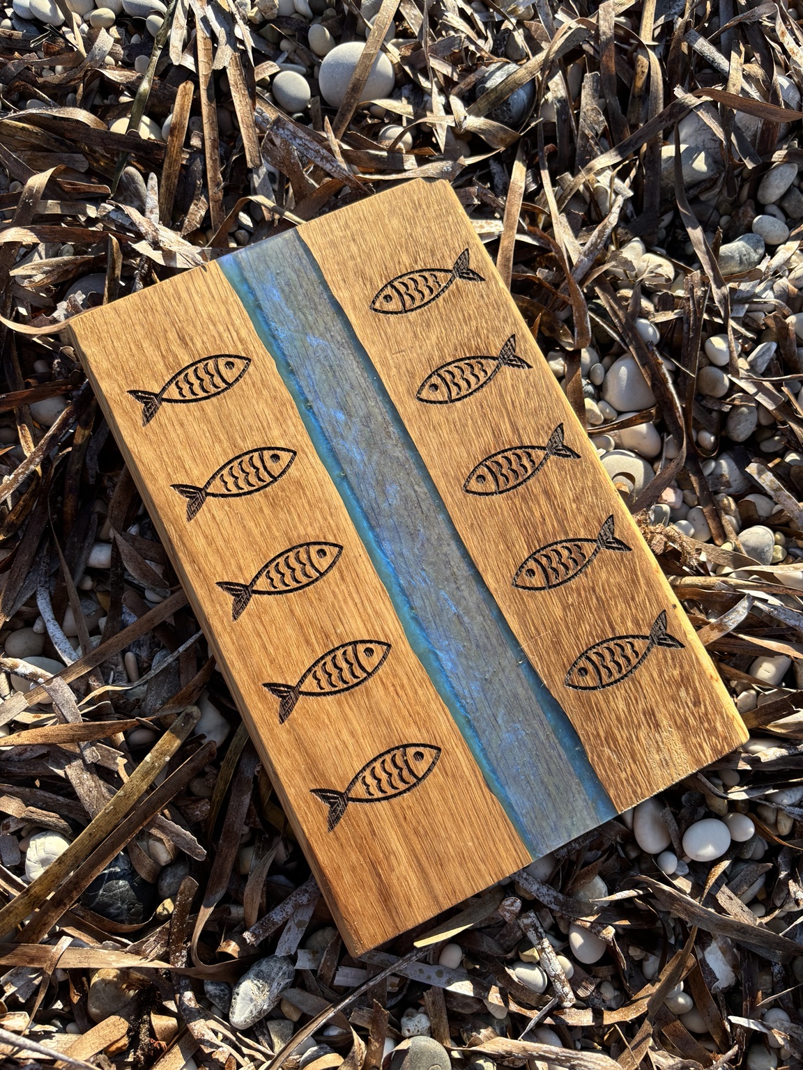 Planche gravée de poissons avec une rivière en résine époxy posée sur une plage de galets